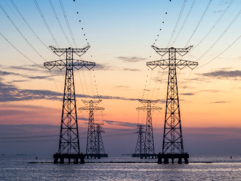 Large metal pylons carry power lines over water, silhouetted against a dramatic sunset of blue to orange.